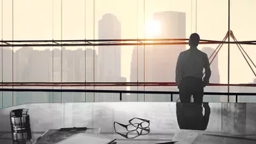 Person looking at city skyline through large office window, desk in foreground.