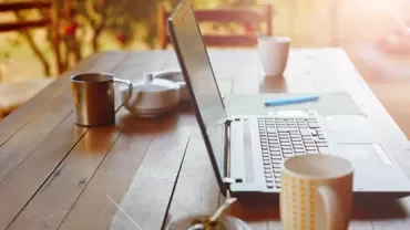 Open laptop on a wooden table with mugs and a teapot in a sunlit setting.