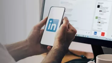 Hands holding a smartphone displaying the LinkedIn logo, with a computer screen in the background.